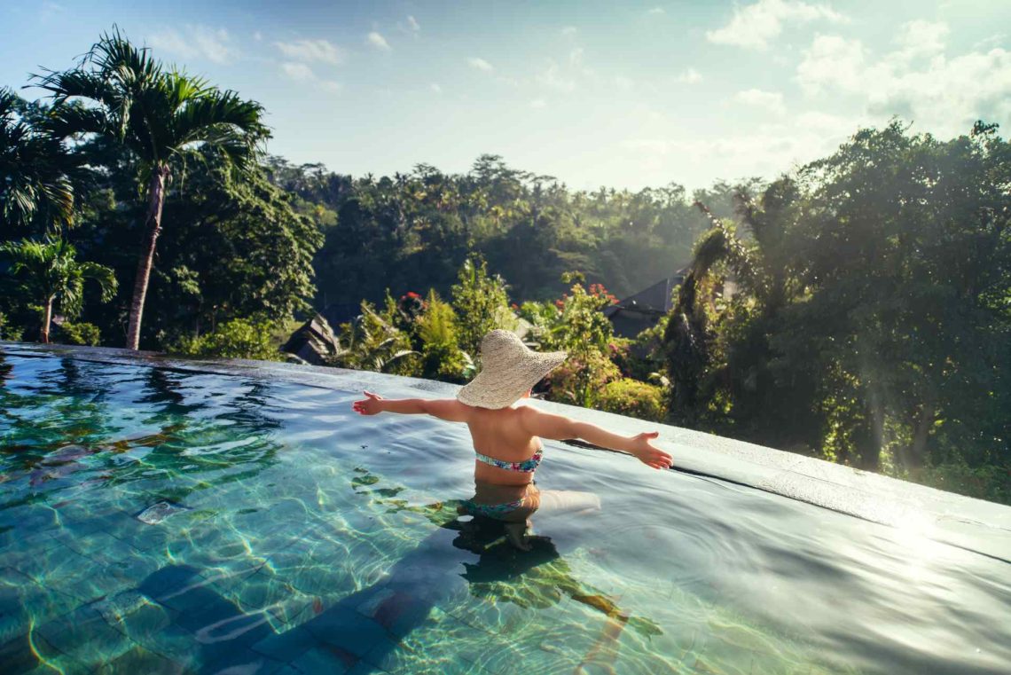 una mujer en una piscina con los brazos abiertos, al fondo mucha naturaleza con palmeras y arboles en dia soleado
