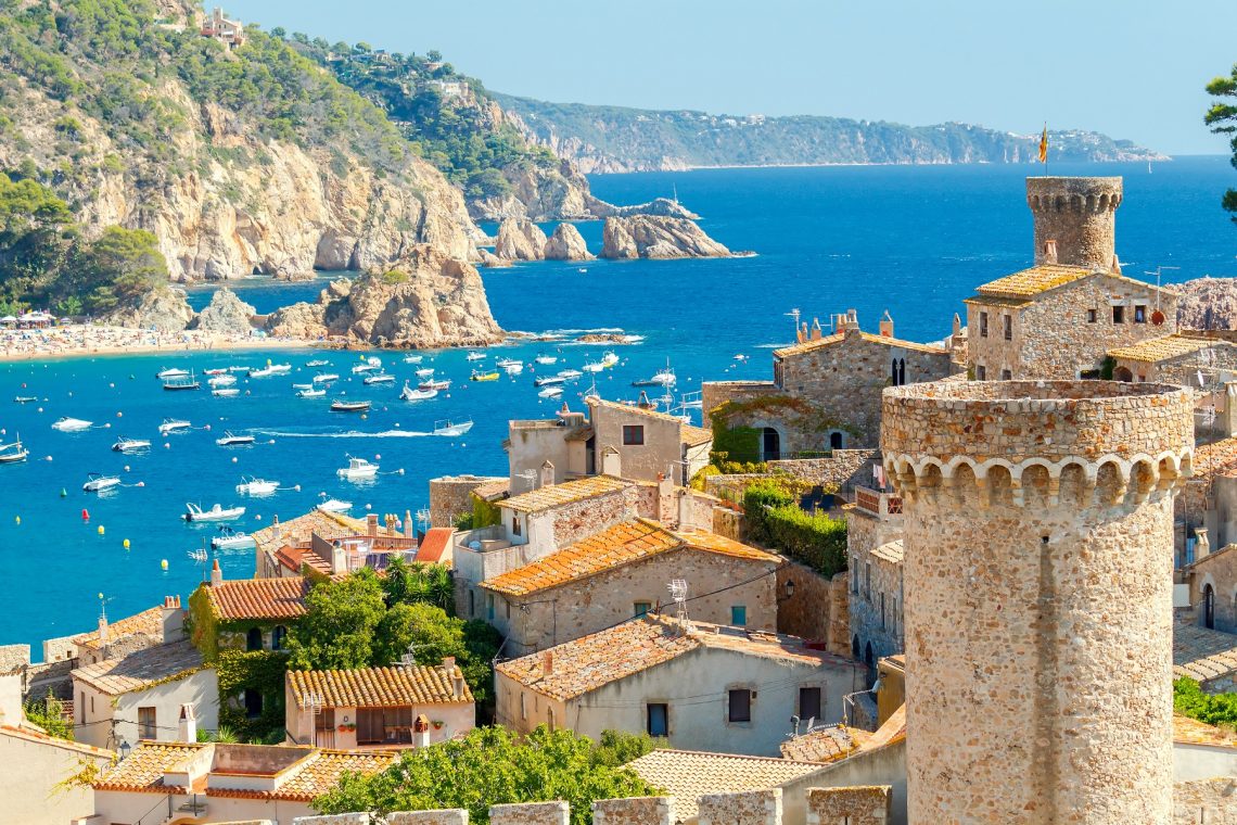 Costa Brava, imagen de pueblo de piedra con barcos al fondo en la bahía, en día de verano